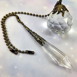 May include: A clear crystal pendulum with a bronze chain and a decorative bronze top. The pendulum is hanging from a clear crystal ball with a bronze top.
