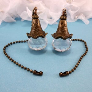 May include: Two antique brass and crystal light pull chains with a decorative floral design. Each chain has a clear crystal ball and a decorative brass finial.