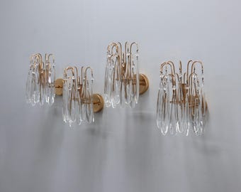 Peut inclure: Quatre appliques murales dorées avec des accents de cristal clair en forme de goutte. Chaque applique a une base circulaire et un cadre incurvé et ouvert contenant les cristaux. Les appliques sont montées sur un mur gris clair.