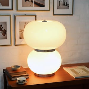 May include: A modern table lamp with a double globe design. The lamp features two spherical, opaque white glass shades stacked on top of each other, separated by a silver band. The lamp sits on a wooden table with books, a small bowl, and a teacup.