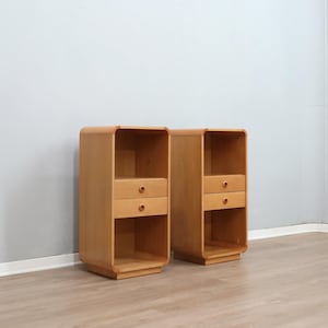 Peut inclure: Deux tables de chevet en bois clair aux bords arrondis. Chaque table de chevet comporte une étagère supérieure ouverte, deux petits tiroirs et une étagère inférieure ouverte. Les tables sont côte à côte.