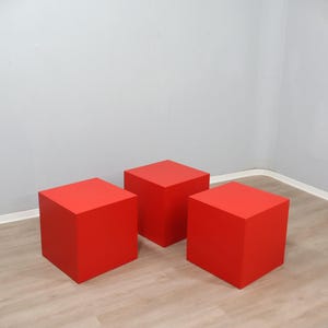 May include: Three bright red square display cubes. The cubes are evenly spaced on a light wood-grain floor, with a neutral gray wall in the background. The cubes are solid and have a matte finish.