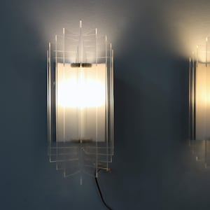 Puede incluir: Dos apliques de pared con un diseño geométrico transparente. Cada aplique tiene un panel rectangular de vidrio esmerilado que emite una luz cálida. Los apliques están montados en una pared azul, con un cable de alimentación negro visible.