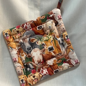 May include: A colorful pot holder with a patchwork design featuring various farm animals, including cows, sheep, goats, and dogs. The pot holder is made of fabric and has a loop for hanging.