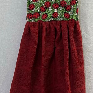 May include: Red kitchen towel with a ladybug print hanging on a white rod. The towel has a red terry cloth body and a green and white ladybug print top.