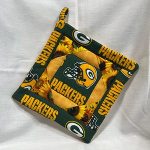 May include: A square, quilted pot holder featuring a green and yellow design. The center displays a football helmet and the word "PACKERS". The border is green with the word "PACKERS" and the team logo. Sunflowers are also featured.