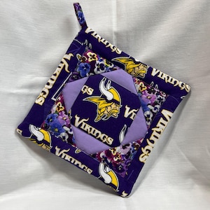 May include: A square purple and lavender pot holder with a loop for hanging. The pot holder features the word "Vikings" and a Viking logo in yellow and white. Floral patterns and a quilted design complete the look.
