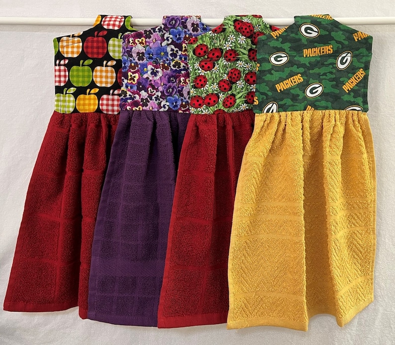 May include: Four kitchen towels hanging on a rack. The towels are red, purple, red, and yellow. The towels have different patterns on them, including apples, flowers, ladybugs, and the Green Bay Packers logo.