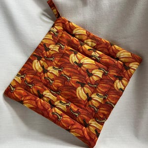 May include: A quilted square pot holder with a repeating pattern of orange and yellow pumpkins. The pot holder has a loop for hanging and is designed for kitchen use. The fabric has a fall theme.