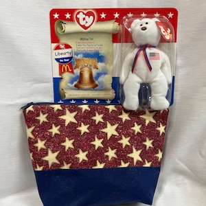 May include: A white plush bear toy with a small American flag patch and red and blue ribbon, packaged with a patriotic themed card. Below is a red, white, and blue star-patterned cosmetic bag.