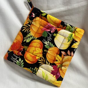 May include: A quilted pot holder with a black background featuring orange pumpkins, fall leaves, and the words "Blessed" and "Grateful" in white script. The pot holder has a loop for hanging.
