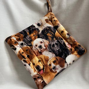 May include: Square quilted pot holder featuring a collage of various dog breeds. The fabric showcases a variety of dogs in shades of brown, black, white, and tan. It has a loop for hanging.
