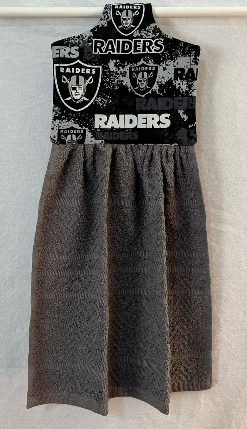 May include: A gray kitchen towel with a black and white pattern featuring the Las Vegas Raiders logo and the word "Raiders".