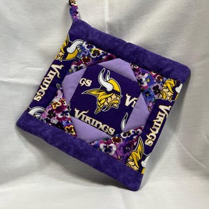 May include: A square, quilted pot holder featuring the Minnesota Vikings logo in purple and gold. The pot holder has a purple border and a hanging loop. The design incorporates floral patterns and the team name.