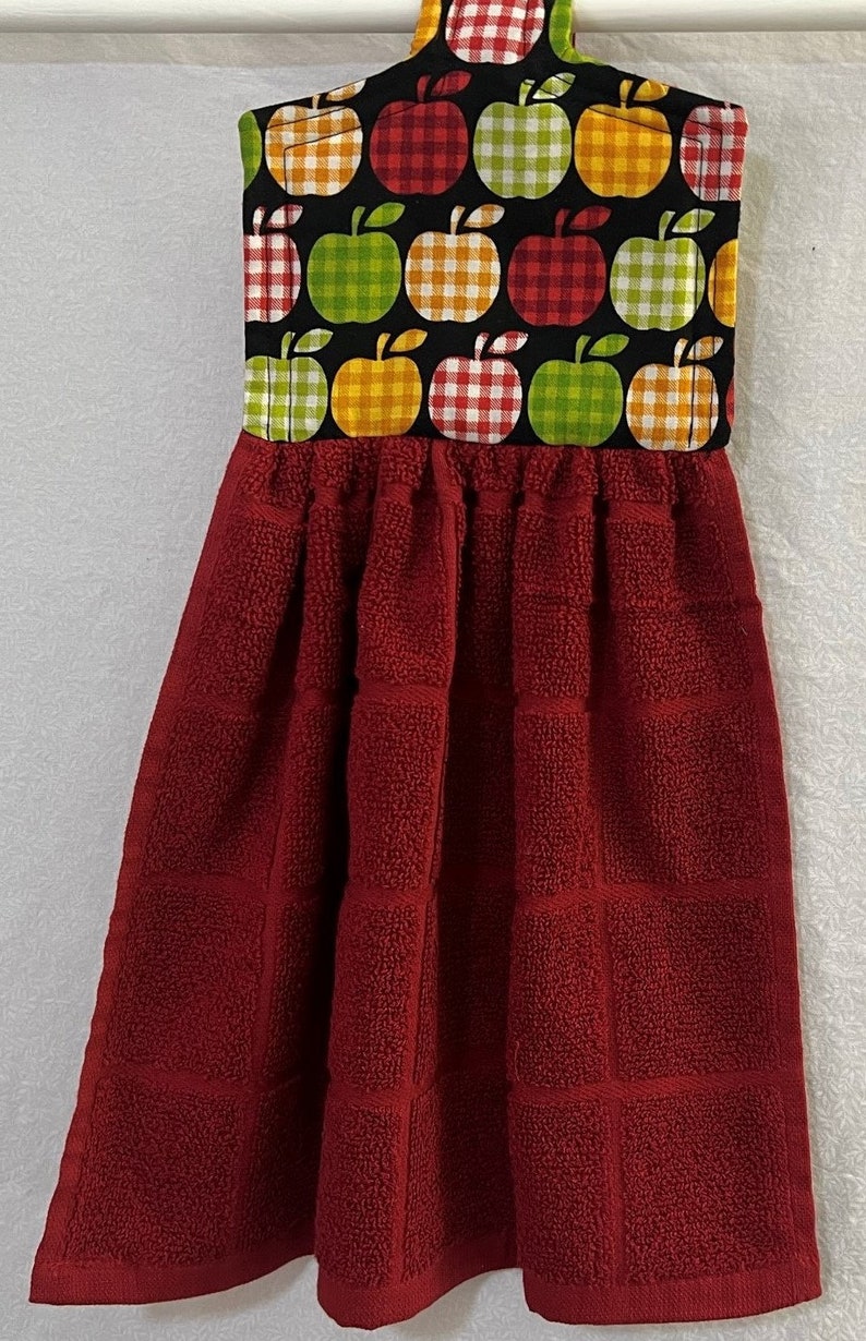 May include: Red kitchen towel with a hanging loop. The top of the towel is decorated with a black fabric with a pattern of green, yellow, and red apples with white gingham.