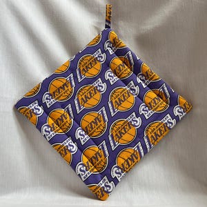 May include: A square purple and gold pot holder featuring a repeating pattern of the Los Angeles Lakers logo. The logo includes a basketball and the text "LOS ANGELES LAKERS". A small fabric loop is attached for hanging.