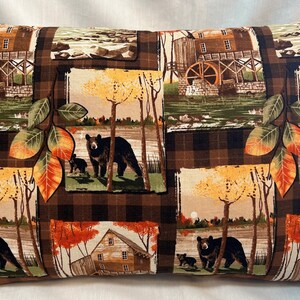 May include: A brown and tan pillow with a plaid pattern and a repeating design of black bears, fall trees, and a gristmill.