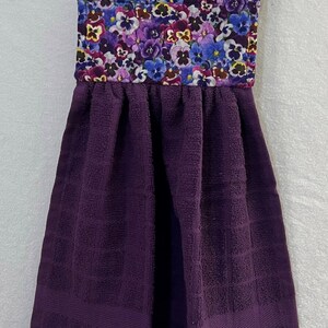 May include: Purple kitchen towel with a floral print hanging loop. The floral print features a variety of purple, blue, and yellow pansies.