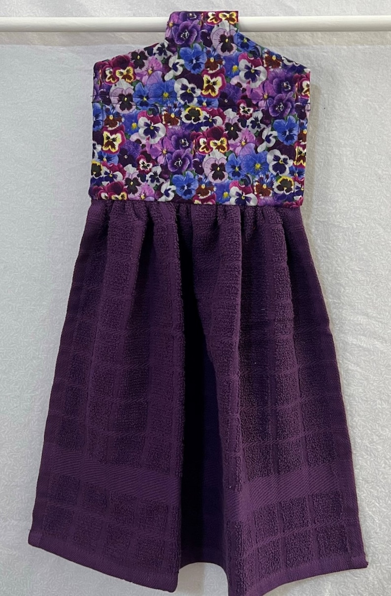 May include: Purple kitchen towel with a floral print hanging loop. The floral print features a variety of purple, blue, and yellow pansies.