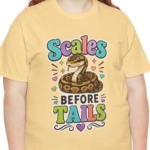 May include: A light yellow t-shirt featuring a graphic of a coiled brown snake. The text above the snake reads "Scales" and the text below reads "BEFORE TAILS" in colorful, bold lettering. Small multi-colored hearts and sparkles surround the snake.