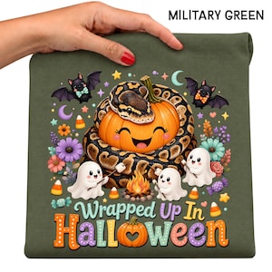May include: A military green t-shirt featuring a graphic of a snake coiled around a smiling pumpkin, two ghosts roasting marshmallows over a campfire, bats, flowers, and candy corn. The text reads "Wrapped Up In Halloween" with a pumpkin icon.