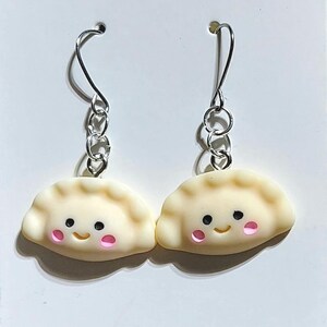 May include: A pair of dumpling-shaped dangle earrings. The charms are a light beige colour with pink cheeks and black eyes. The earrings have silver-coloured hooks and chain links.