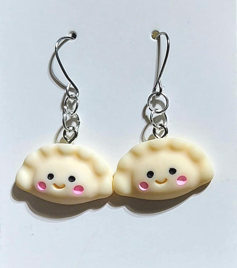 May include: A pair of dumpling-shaped dangle earrings. The charms are a light beige colour with pink cheeks and black eyes. The earrings have silver-coloured hooks and chain links.
