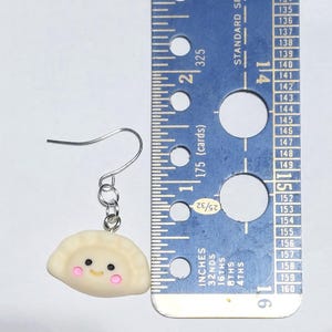 May include: A single dumpling earring with a silver hook. The dumpling is off-white with pink cheeks and black eyes. A blue ruler with cm measurements is next to the earring.