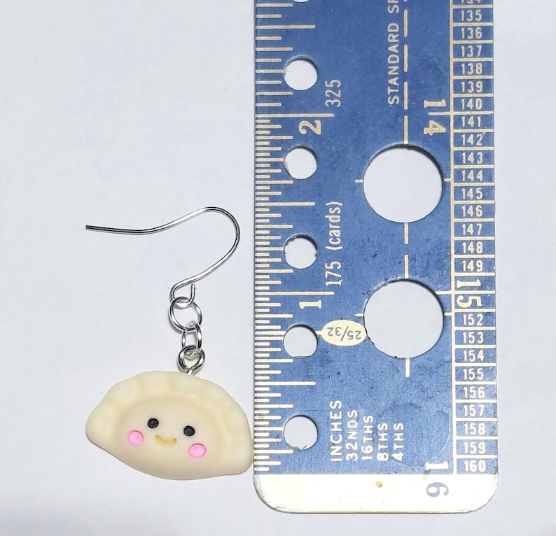 May include: A single dumpling earring with a silver hook. The dumpling is off-white with pink cheeks and black eyes. A blue ruler with cm measurements is next to the earring.
