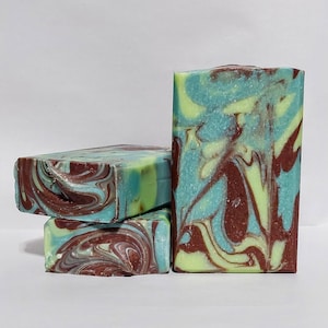 May include: Four handmade soap bars with a marbled design. The soaps feature swirls of teal, brown, and light green. The bars are rectangular and stacked, showcasing the color variations and textures.