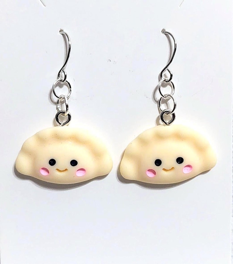 May include: A pair of dumpling-shaped dangle earrings. The charms are light beige with black eyes and pink cheeks. The earrings have silver-coloured fish hook ear wires.