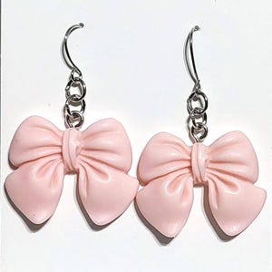 May include: A pair of light pink bow earrings. Each earring features a bow shape with a textured surface, connected to a silver-colored chain and hook. The earrings are displayed against a white background.