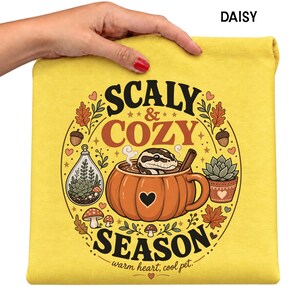 May include: A bright yellow t-shirt featuring a graphic print of a snake inside a pumpkin-shaped mug. The text reads SCALY & COZY SEASON and warm heart, cool pet. The design includes a terrarium, a succulent, mushrooms, acorns, and autumn leaves.