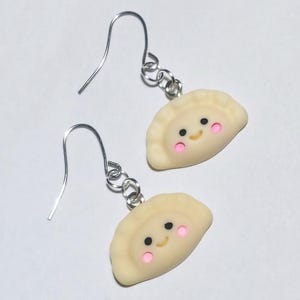 May include: A pair of handmade earrings featuring smiling dumpling charms. The charms are light yellow with pink cheeks and black eyes. The earrings have silver-coloured fish hook ear wires.