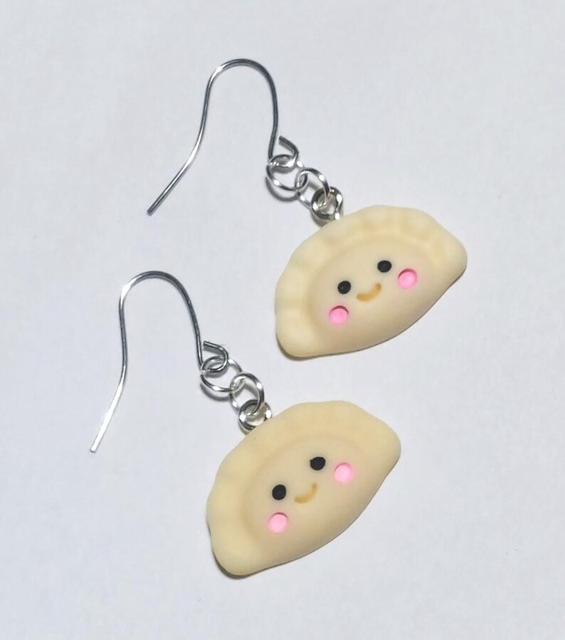 May include: A pair of handmade earrings featuring smiling dumpling charms. The charms are light yellow with pink cheeks and black eyes. The earrings have silver-coloured fish hook ear wires.