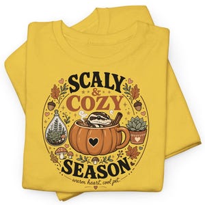 May include: A mustard yellow t-shirt featuring a circular graphic with the text "SCALY & COZY SEASON" and "warm heart, cool pet." The design includes a snake inside a pumpkin mug, a terrarium, a succulent, mushrooms, leaves, and acorns.