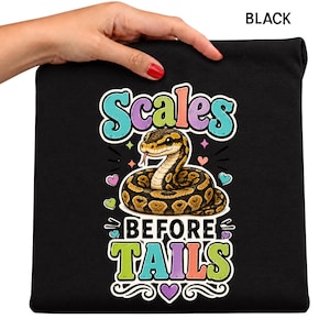 May include: A black t-shirt featuring a graphic of a coiled brown and black snake. The text "Scales BEFORE TAILS" is printed in colorful, bubbly letters above and below the snake, surrounded by small heart and star icons on the black fabric.