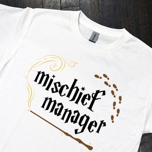 May include: White t-shirt with a black graphic that says "mischief manager" with a brown wand and gold sparkles. There are brown footprints around the text.