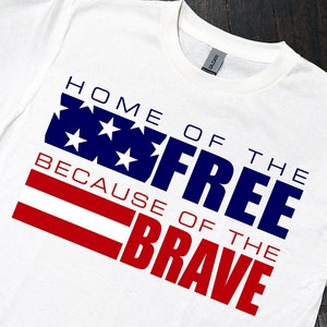Op de afbeelding: Wit t-shirt met de tekst "HOME OF THE FREE BECAUSE OF THE BRAVE" in blauw en rood. Het ontwerp bevat een blauwe rechthoek met witte sterren en rode strepen, die op een vlag lijken.