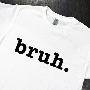 May include: White t-shirt with the word "bruh" printed in black bold letters.