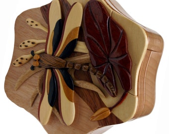 May include: A wooden puzzle box with a dragonfly and leaf design. The dragonfly has brown, black, and tan wings. The leaf is a deep red color. The box is made of various wood tones and has a unique shape.