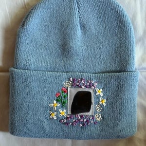 May include: A light blue knit beanie with a folded brim. The beanie features an embroidered floral design with a square patch in the center. The floral design includes white, yellow, red, and purple flowers.