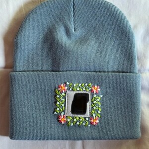 May include: A light blue beanie with a square embroidered patch. The patch features a floral design with pink and yellow flowers, green leaves, and small white accents. The beanie is made of a soft, knitted material.