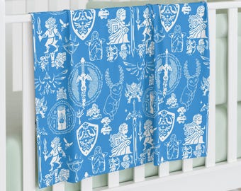 May include: A blue baby blanket with a white pattern of various symbols and characters from the Legend of Zelda video game series. The pattern includes the Triforce, Master Sword, and other iconic elements.