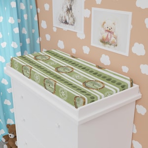 May include: A green and white changing pad cover with a repeating pattern of round doors and vertical stripes. The cover is placed on top of a white changing table. The nursery decor includes framed teddy bear art.