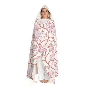 May include: A hooded blanket with a white background and a pink floral pattern featuring stylized creatures. The blanket is made of soft, plush fabric and is perfect for snuggling up on the couch.