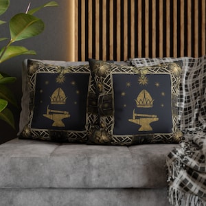May include: Two decorative throw pillows with a dark blue background and gold accents. The pillows feature a stylized design of a hammer and anvil with a crown on top. The design is surrounded by a gold frame with intricate details.