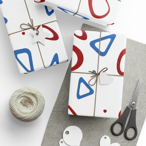 Pu&ograve; includere: Tre scatole regalo bianche con motivi geometrici rossi e blu. Le scatole sono avvolte con spago e hanno etichette di carta bianca attaccate.