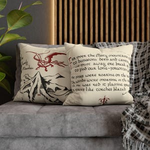 Op de afbeelding: Twee decoratieve sierkussens op een grijze bank. Eén kussen heeft een rode draak en berg illustratie met de tekst "The Lonely Mountain". Het andere kussen heeft handgeschreven tekst in zwarte en rode inkt.