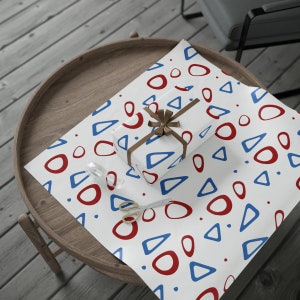 Pu&ograve; includere: Una scatola regalo bianca avvolta in carta con motivo geometrico rosso e blu. La carta presenta un motivo ripetitivo di cerchi e triangoli. Un nastro marrone &egrave; legato attorno alla scatola con un fiocco.